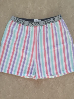 Shorts - Färgglada shorts från Wow med vertikala ränder i lila, grön, röd, blå och vit. De har en bred resår i midjan med logotyp och är tillverkade i mjuk bomull. Passformen är loose och shortsen är perfekta för sommaren.