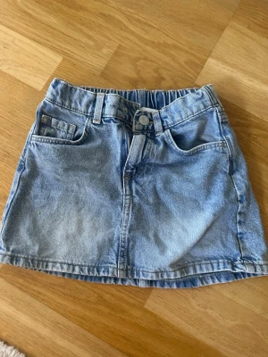 Blå jeanskjol med fickor - Snygg blå jeanskjol i klassisk modell med fram- och bakfickor samt hällor för bälte. Kjolen har knapp och dragkedja framtill och resår i midjan baktill för extra komfort. Storlek 116.
