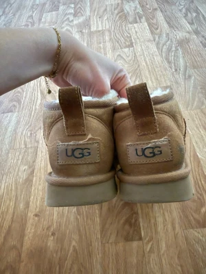 UGG mini - Säljer ett par klassiska bruna UGG boots i mocka med mjukt vitt fårskinnsfoder. Skorna har en låg modell och bred passform, samt dragflikar bak för enkel påtagning. Perfekta för kalla dagar när du vill ha både stil och komfort. Finns defekter som syns på bilderna 