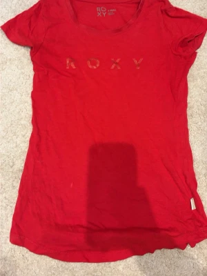 Röd t-shirt från Roxy - Snygg röd t-shirt från Roxy med rund halsringning och diskret logga framtill. Tillverkad i mjuk bomull och har en klassisk passform med korta ärmar. Perfekt för en avslappnad och cool stil. Liten fläck 