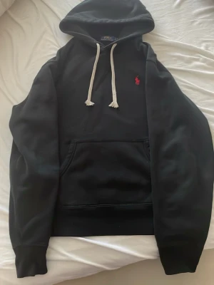 Svart hoodie från Polo Ralph Lauren - Svart hoodie från Polo Ralph Lauren med vit dragsko i huvan och röd broderad logga på bröstet. Klassisk känguruficka framtill och ribbade muddar vid ärmar och midja. Tillverkad i mjukt bomullsmaterial för en skön och avslappnad look.