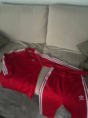 Röd Adidas SST - Rött set från Adidas med klassiska vita ränder längs ärmar och ben. Setet består av en zip-jacka och matchande byxor, båda med Adidas logga framtill. 