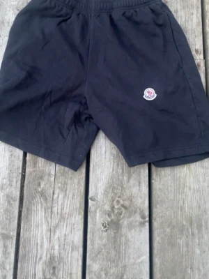 Svarta shorts från Moncler i bomull - Svarta shorts från Moncler med elastisk midja och klassisk logotyp på ena benet. Tillverkade i mjuk bomull, perfekta för en avslappnad stil. Passar dig som gillar stilrena och bekväma plagg.