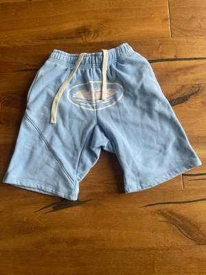 Ljusblåa corteiz shorts - Säljer ett par ljusblåa shorts med bred passform och vit snörning i midjan. Shortsen har ett tryck framtill och en bakficka. Perfekta för chill dagar och sommarhäng.