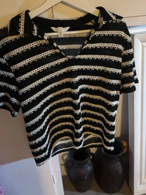 Randig svartvit topp från H&M - Svart och vit randig topp från H&M med kort ärm och v-ringning. Toppen har virkade detaljer i de vita ränderna och en avslappnad passform. Perfekt för en trendig och chill look.