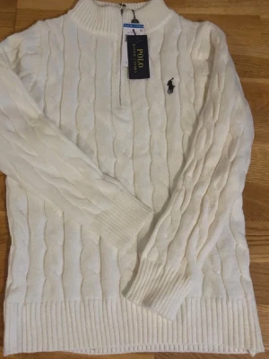 Vit kabelstickad tröja Polo Ralph Lauren - Kabelstickad vit tröja från Polo Ralph Lauren med klassisk svart logga broderad på bröstet. Tröjan har rund halsringning, långa ärmar och ribbade muddar vid ärmslut och nederkant. Perfekt för en stilren och tidlös look.
