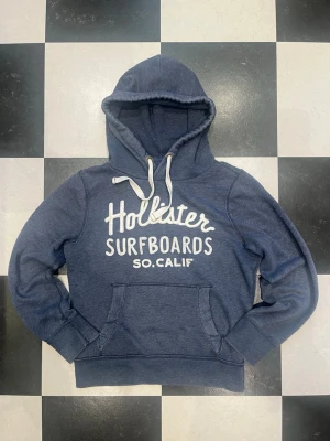 Hollister hoodie - Mysig blå hoodie från Hollister med vit text 'Hollister Surfboards SO.CALIF' på bröstet. Hoodien har en stor huva med vita dragsnören och en klassisk magficka framtill. Perfekt för en avslappnad och cool stil.