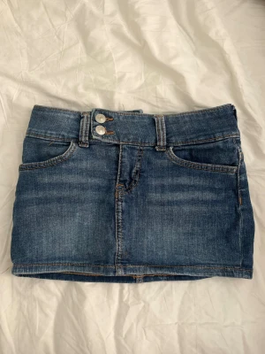 Lågmidjad kjol från hm - Söt blå jeanskjol med två knappar framtill och klassiska fickor både fram och bak. På ena bakfickan finns en liten rosa broderad hjärtdetalj. Kjolen har bälteshällor och en kort, rak passform som ger en trendig look.