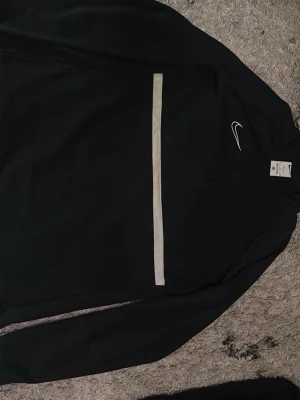 Svart Nike Dri-Fit träningsjacka - Säljer en svart träningsjacka från Nike med Dri-Fit material som transporterar bort fukt. Jackan har en vit horisontell rand på bröstet och en liten vit Nike-logga på vänster sida. Perfekt för träning eller sportiga dagar.