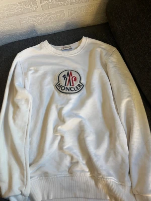 Vit sweatshirt från Moncler - Snygg vit sweatshirt från Moncler med stort broderat logotypmärke på bröstet i rött, blått och vitt. Tröjan har rund halsringning och ribbade muddar vid ärmslut och nederkant. Perfekt för en clean och stilren look.