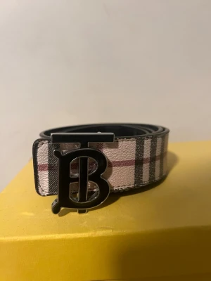 Rutig Burberry bälte med B-spänne - Snyggt bälte från Burberry med klassiskt rutigt mönster i beige, svart och rött. Bältet har ett stort metallspänne med Burberrys ikoniska B-logga. Perfekt accessoar för att lyfta din outfit.