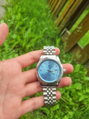 Silverfärgad klocka från Ricecgo - Stilren klocka från Ricecgo med silverfärgat metallarmband och ljusblå urtavla. Urtavlan har arabiska siffror och datumvisning. Klassisk design med räfflad bezel och länkarmband som ger en modern vibe. Helt ny aldrig använd och ej kortad så alla länkar ingår