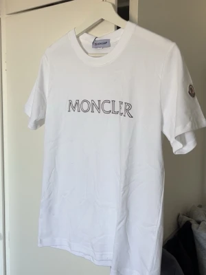 Vit Moncler t-shirt med logga - Snygg vit t-shirt från Moncler med klassisk logga broderad på ärmen och tryckt Moncler-text på bröstet. Tshirten är true to size så att om du har s på alla fina kläder så kommer tshirten sitta perfekt. T-shirten har rund hals och korta ärmar. Tillverkad i mjuk bomull för en skön känsla och clean look. PRIS GÅR ATT DISKUTERAS!
