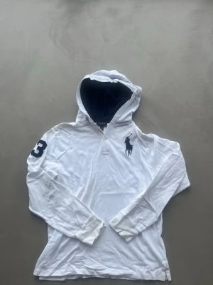Vit hoodie från Polo Ralph Lauren - Vit hoodie från Polo Ralph Lauren med stor marinblå logga på bröstet och siffran 3 på ena ärmen. Hoodien har långa ärmar, huva med mörkblå insida och är tillverkad i mjukt bomullstyg. Perfekt för en avslappnad och sportig stil.