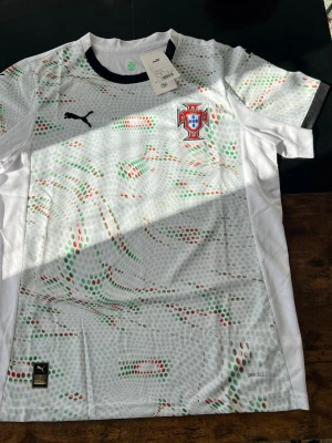 Portugal fotbollströja Puma vit puma protugal football jersey  - Snygg vit Portugal fotbollströja från Puma med färgglada prickmönster i grönt och rött. Tröjan har Portugal-emblemet på bröstet och Puma-logga. Tillverkad i lätt och ventilerande material, perfekt för träning eller match. WITH RECEIPT 