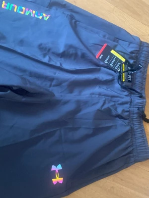 Blå träningsshorts från Under Armour - Snygga blå träningsshorts från Under Armour med regnbågsfärgad logga och tryck. Shortsen har elastisk midja med snörning och är gjorda i ett lätt och snabbtorkande material, perfekta för träning eller sport.