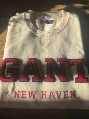 Vit college-tröja från GANT - Snygg vit college-tröja från GANT med stora röda bokstäver och texten 'NEW HAVEN' broderat framtill. Tröjan har rund halsringning, ribbade muddar och är tillverkad i mjukt material. Perfekt för en avslappnad och trendig look. Skriv om du har frågor eller vill ge bud!