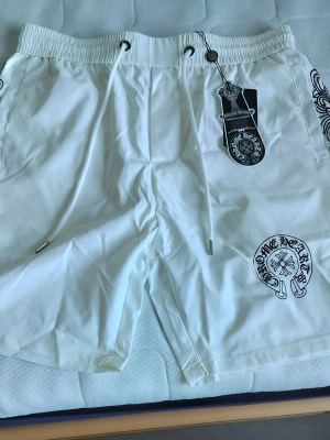 Vita shorts med tryck från Chrome Hearts - Snygga vita shorts från Chrome Hearts med svart tryckdetalj på ena benet och sidan. Shortsen har snörning i midjan och elastisk linning för skön passform. Perfekta för sommaren och har en cool streetwear-vibe.