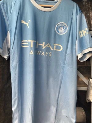 Manchester City matchtröja Puma - Snygg ljusblå Manchester City matchtröja från Puma med klubbmärke och Etihad Airways-tryck på bröstet. Nummer 10 och namnet Cherki på ryggen. Tillverkad i lätt och ventilerande polyester, perfekt för fotboll eller träning.