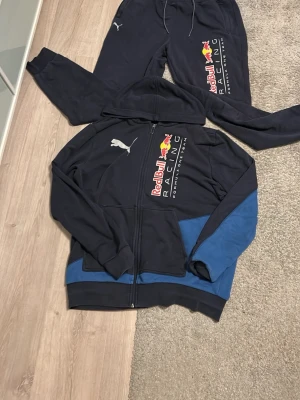 Red Bull Racing hoodie från Puma - Mörkblå hoodie från Puma med Red Bull Racing-logga och dragkedja framtill. Hoodien har huva, fickor och blå detaljer på ärmar och nederkant. Tillverkad i mjukt bomullsmaterial, perfekt för dig som gillar motorsport och streetwear.