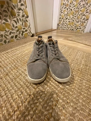 Gråa Louboutins - Säljer ett par stilrena gråa sneakers i mocka med röda sulor och klassisk snörning. Skorna har en låg profil och diskret design som passar till det mesta. Insidan är lätt vadderad för extra komfort.