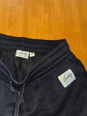 Svarta mjukisbyxor från Andy XS - Svarta mjukisbyxor från Andy med dragsko i midjan och ribbade muddar vid bensluten. Byxorna har en relaxed fit och en liten logga på vänster sida. Perfekta för chill dagar eller när du vill vara extra bekväm.