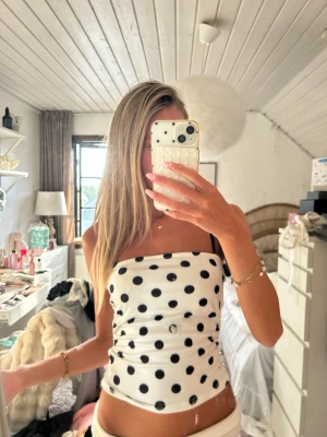 Vit prickig tubtopp med svarta detaljer - Snygg vit tubtopp med svarta prickar och tunna axelband. Toppen har en rak bandeau-form och är perfekt för varma dagar. Materialet känns mjukt och stretchigt, vilket gör den bekväm att bära. Enkel men trendig design som sticker ut.