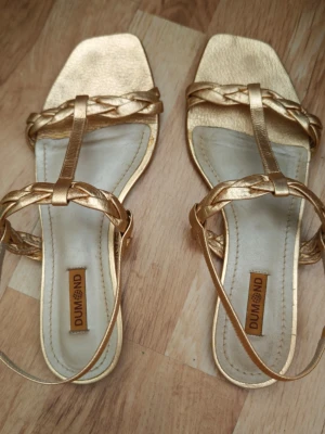Guldfärgade sandaler Dumond - Eleganta guldfärgade sandaler från kvalitetsmärket Dumond. Perfekta till sommarens fester, bröllop, student, semester eller när du vill piffa upp en vardagsoutfit. Sandalerna har en stilren design med flätade remmar och justerbar ankelrem som ger en bekväm passform. Använda få gånger och i mycket fint skick. Normalt slitage på sulorna, men ovandelen är välbevarad.  Märke: Dumond Storlek: 39 (EU) Färg: Guld Justerbar ankelrem med spänne Bekväma och lätta att bära Tillverkade i Brasili