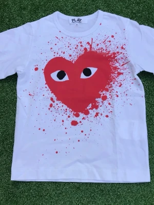 Vit t-shirt PLAY med rött hjärta - Vit t-shirt från PLAY Comme des Garçons med stort rött hjärta och ögon på bröstet, omgiven av röda färgstänk. Klassisk rund hals och korta ärmar. Perfekt statement-plagg för dig som gillar streetstyle.
