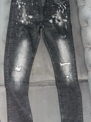Grå slitna jeans Surly & Demand - Säljer ett par grå jeans från Surly & Demand med skinny fit och slitna detaljer samt färgstänk. Jeansen har en tvättad look och lagade partier på benen för en trendig streetstil. Perfekta för dig som gillar en edgy vibe.