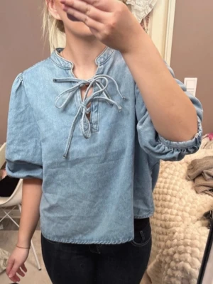 Ljusblå jeansblus med puffärm - Trendig ljusblå jeansblus med puffiga trekvartsärmar och snygg knytning vid halsen. Blusen har en avslappnad passform och är gjord i mjukt denimtyg. Perfekt att styla med både jeans och kjol för en cool look.