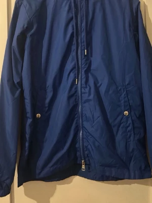 Blå vindjacka från Moncler - Snygg blå vindjacka från Moncler med dragkedja framtill och två fickor med tryckknappar. Jackan har Moncler-logga på ärmen och är tillverkad i ett lätt, vindtätt material. Perfekt för blåsiga dagar och enkel att matcha med olika outfits.