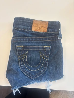 Blå jeansshorts från True Religion - Säljer ett par blå lågmidjade jeansshorts från True Religion med råa, fransiga kanter och slitna detaljer. Shortsen har klassiska fickor fram och bak, samt den ikoniska True Religion-sömmen på bakfickan. Perfekta för sommaren och har en cool, avslappnad vibe. W25 är storleken, jag skulle säga att dom är mindre i storlek.