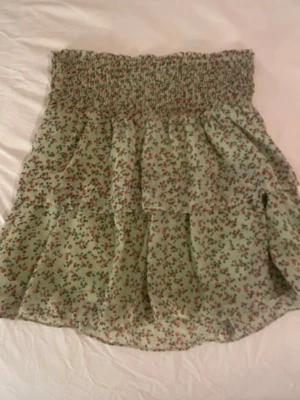 Grön blommig volangkjol - Söt kort grön kjol med småblommigt mönster i rött och rosa. Kjolen har bred smock i midjan och volangdetaljer som ger en luftig och somrig känsla. Perfekt för dig som gillar romantisk och avslappnad stil.