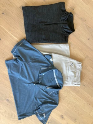 Golfkläder Puma och J.Lindeberg - Snygg mörkgrå tröja från J.Lindeberg med half zip-dragkedja framtill. Tillverkad i ett mjukt och stretchigt syntetmaterial som passar perfekt för en sportig och modern look. Storlek M. Ljusgråa Puma-byxor i storlek 30/30. Ljusblå Puma-piketröja i mjukt material i storlek XS. 