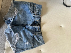 Blå lågmidjade shorts från 157 - Snygga blå denimshorts från 157 med slitna detaljer och råa kanter. Klassisk femficksmodell med bälteshällor och knappgylf. Perfekta för sommaren och ger en avslappnad vibe.