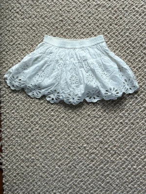 Vit kortkjol med broderade blommor - Supersöt vit kortkjol med elastisk midja och broderade blommor längs hela kjolkanten. Kjolen har en luftig och volangig form som ger en söt vibe. Perfekt för varma dagar och somriga outfits!