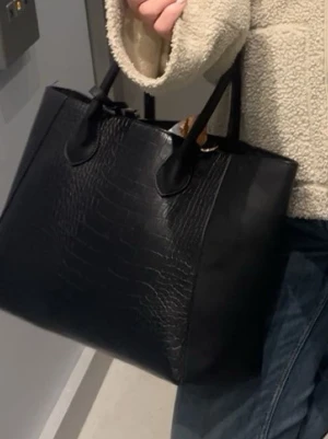 Svart stor väska  - Stilren svart shoppingväska med krokodilpräglat mönster och dubbla handtag. Väskan är rymlig och har en klassisk fyrkantig form, perfekt för att bära med sig allt man behöver. Materialet ser ut att vara konstläder.