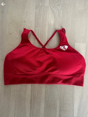 Röd sport-bh med korsade band - Snygg röd sport-bh med korsade axelband i ryggen och bred resår under bysten. Bh:n har en liten vit logga framtill och är tillverkad i ett stretchigt, mjukt material som ger bra stöd. Perfekt för träning eller en aktiv vardag.