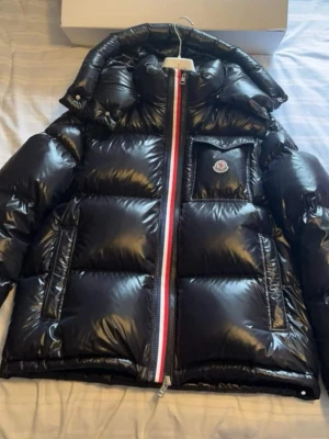 Svart dunjacka från Moncler - Säljer en svart, glansig dunjacka från Moncler med avtagbar huva och klassisk logotyp på bröstet. Jackan har en vit, röd och blå dragkedja samt två sidofickor med dragkedja. Perfekt för kalla vinterdagar och riktigt snygg att bära.