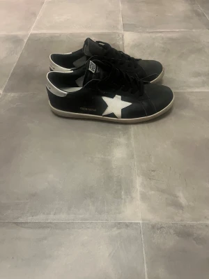 Svarta Golden Goose Super-Star sneakers - Säljer ett par svarta Golden Goose Super-Star sneakers med vita detaljer och klassisk GGDB-logga på tungan. Skorna har en låg profil, svarta skosnören och vit sula. Materialet är läder och textil, perfekt för dig som gillar stilrena och trendiga sneakers.