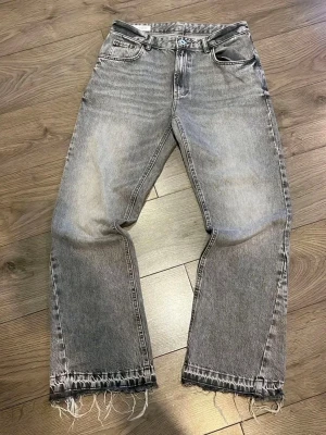 Grå jeans med råa kanter - Säljer ett par grå jeans med vintage tvätt och råa, fransiga kanter längst ner. Jeansen har klassisk femficksdesign och raka ben. Perfekt för dig som gillar en avslappnad och trendig look. Materialet är denim.