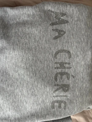 Grå hoodie med texten MA CHÉRIE - Säljer en ljusgrå hoodie med texten 'MA CHÉRIE' i glittriga stenar på bröstet. Hoodien har huva med dragsko och är tillverkad i mjukt sweatshirtmaterial. Perfekt för dig som gillar en enkel men ändå snygg stil.