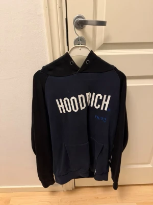 Svart och marinblå HOODRICH hoodie - Snygg hoodie från HOODRICH i svart och marinblått med stor vit logga på bröstet. Klassisk känguruficka framtill och huva med dragsko. Perfekt för en avslappnad streetwear-look. Tillverkad i mjukt bomullsmaterial.