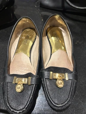 Svarta loafers från Michael Kors - Snygga svarta loafers från Michael Kors i läder med guldfärgade detaljer och MK-logga framtill. Skorna har en klassisk form och guldfärgad innersula som ger en lyxig känsla. Perfekta för dig som gillar stilrena och exklusiva skor.