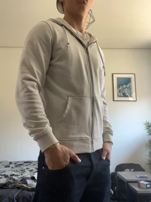 Ljusgrå zip-hoodie med huva - En stilren ljusgrå hoodie med dragkedja och huva. Modellen har ribbade muddar, två stora fickor framtill och justerbara snören i huvan. Perfekt för en avslappnad och enkel look. Materialet känns mjukt och bekvämt mot huden.