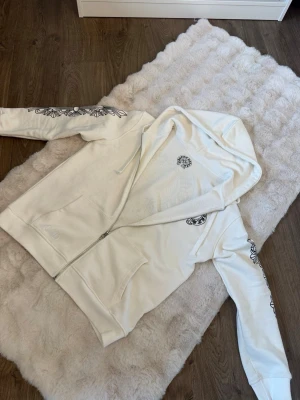 Chrome hearts zip up  - Snygg vit hoodie med dragkedja från Chrome Hearts. Hoodien har svarta tryck på båda ärmarna och ett tryck på bröstet. Utrustad med huva och dragsnören samt två fickor framtill. Perfekt för en streetwear-look. Aldrig använt tag följer med