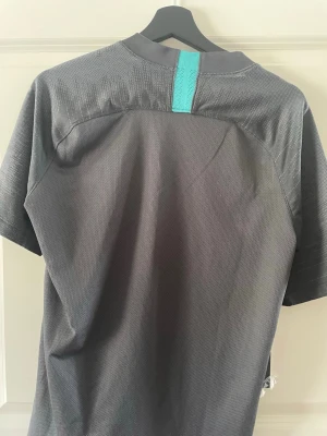 FC Barcelona tränings t-shirt Nike - Mörkgrå FC Barcelona tränings t-shirt från Nike med klubbmärke och turkos Nike-logga på bröstet. Beko sponsortryck framtill. Tillverkad i ett lätt och ventilerande funktionsmaterial, perfekt för träning eller sport.