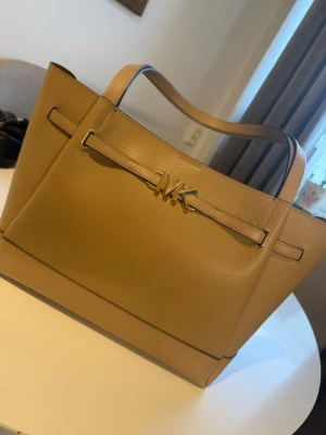 Beige handväska från Michael Kors - Stilren beige handväska från Michael Kors i slätt och lätt strukturerat läder. Väskan har två rejäla handtag, guldfärgad MK-logga framtill och en rymlig, fyrkantig form. Perfekt för dig som gillar klassisk och enkel design med en touch av lyx.