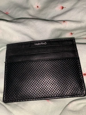 Svart kortfodral från Mario Valentino - Stilrent svart kortfodral från Mario Valentino i äkta läder med perforerat mönster på framsidan. Fodralet har flera kortfack och en diskret logga upptill. Perfekt för dig som vill ha en smidig och snygg plånbok i fickan.